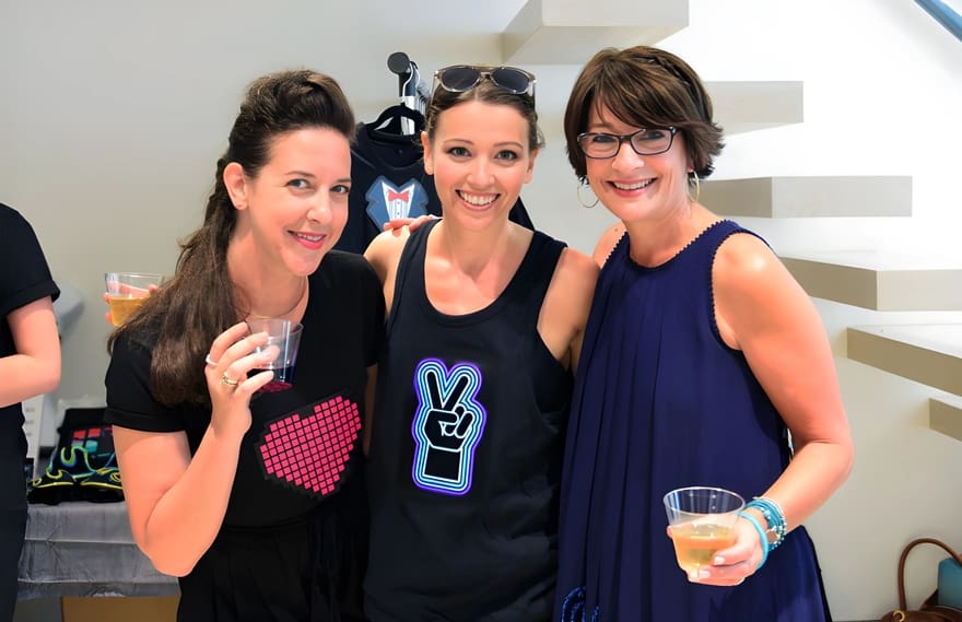 Three women pose for a photo, two wearing light-up t-shirts, each holding a drink and smiling