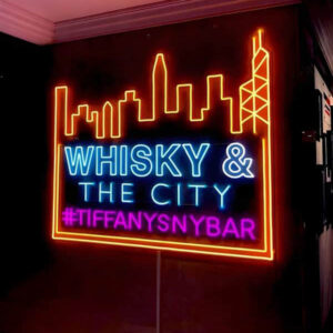 Neon sign for bar with NYC city skyline lighting up in orange and the words Whisky & The city lighting up blue with a purple tagline reading #tiffanysnybar