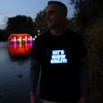 Smiling guy wearing "lets glow crazy" light up shirt outside in front of water and LED swan boats