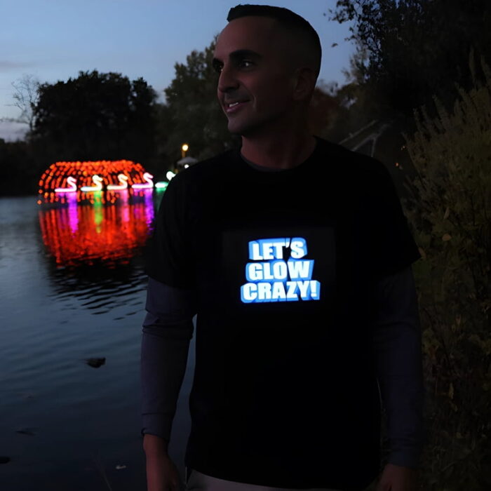 Smiling guy wearing "lets glow crazy" light up shirt outside in front of water and LED swan boats