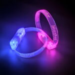 Two bracelets with custom engraved Muster logo lighting up bright blue and hot pink in dark room