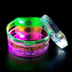 Three custom LED bracelets stacked up with another white LED bracelet leaning against it in a dark room, all glowing in LED colors
