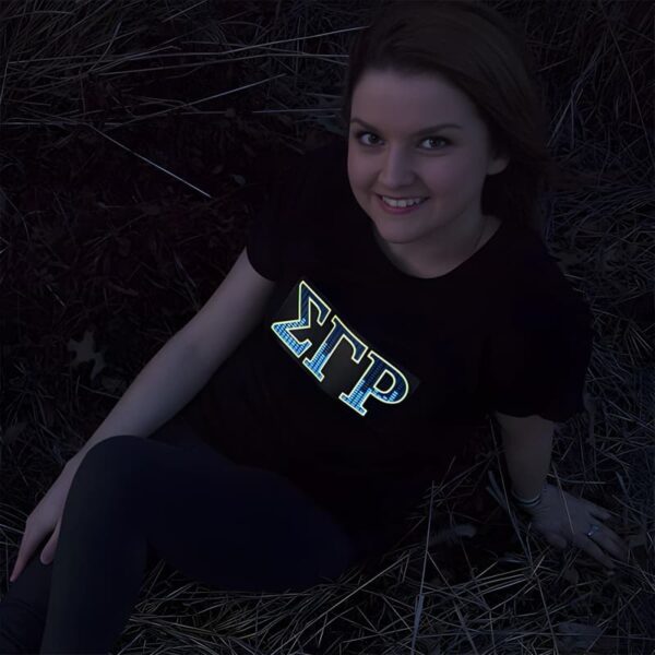 Smiling girl posing in dark while sorority greek letters are lighting up on her shirt