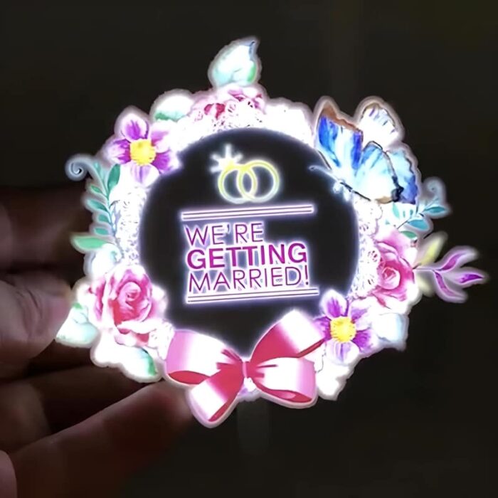 Hand holding custom EL light up panel with we're getting married text, rings, and flower wreath lighting up bright neon colors