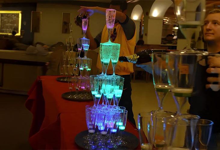 A man pours drinks into glasses at a bar, with illuminated champagne glasses arranged in a tower for a wedding celebration