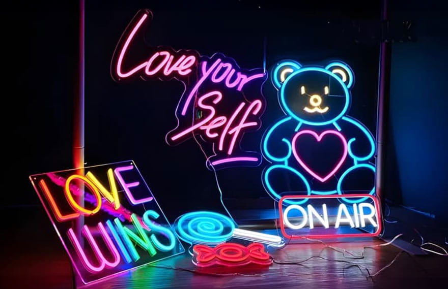 Six different neon signs in dark room with multiple colors lighting up brightly including a blue lollipop, red candy wrapper, on air, teddy bear, a rainbow sign reading "love wins" and a pink sign reading "love your self"