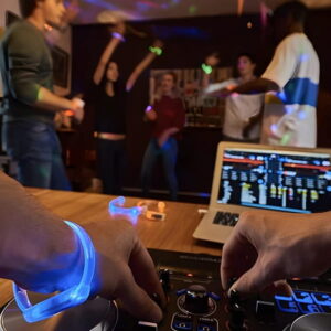 DJ wearing light up bracelets while operating turntable in front of crowd of dancing people wearing different colored light up bracelets