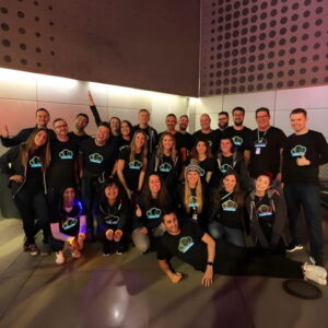 A team of individuals wearing black LED shirts, enthusiastically raising their hands during a tech event