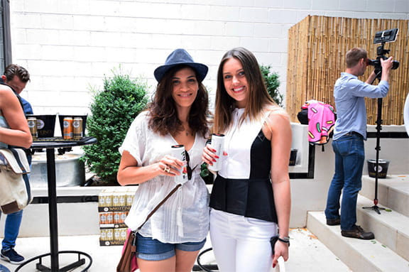 Two women smiling and posing together for a photo at an outdoor event, surrounded by greenery and festive decorations