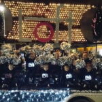 A group of dancers on a float, wearing illuminated t-shirts with their logo, accompanied by a bright banner
